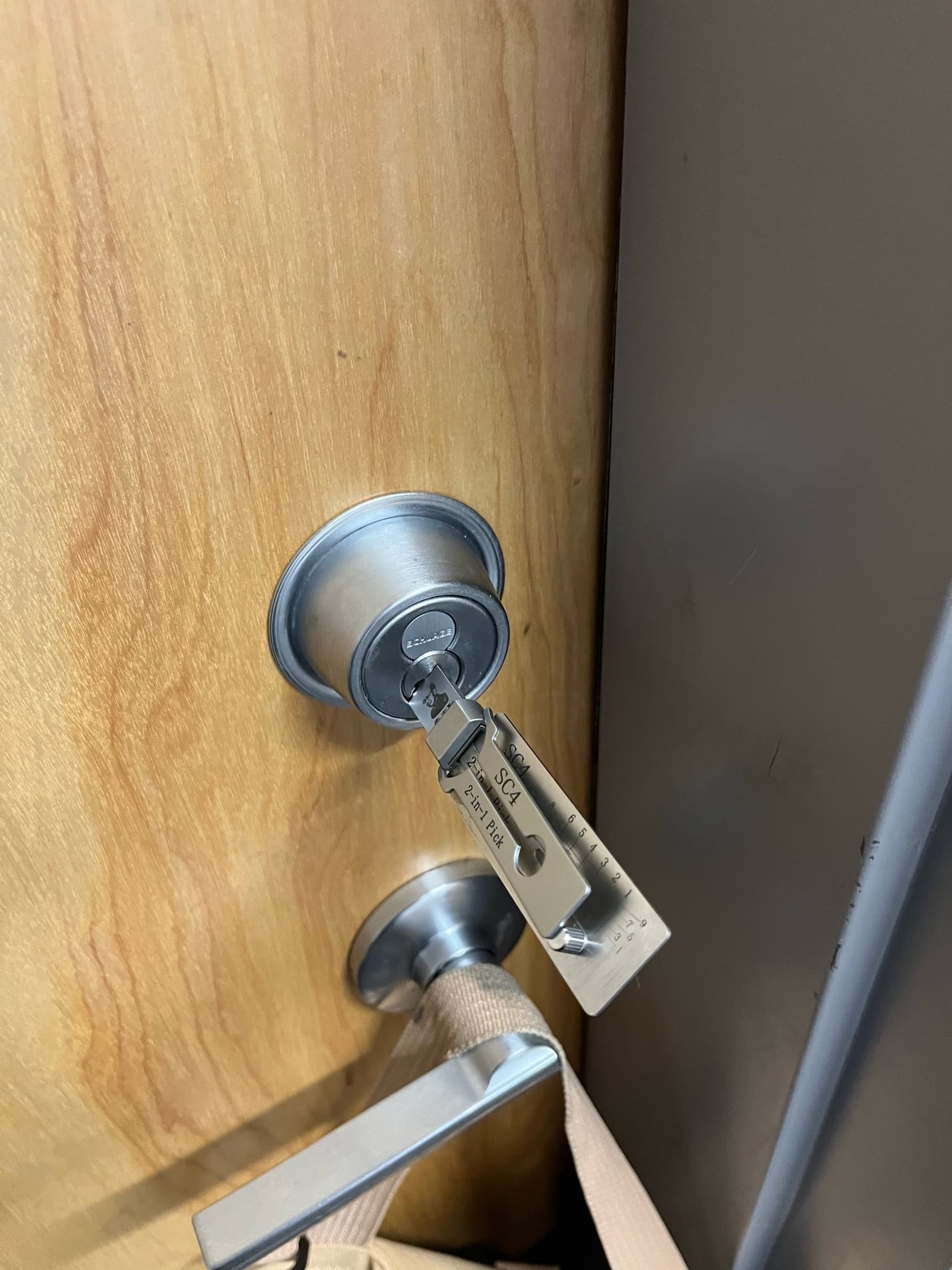 Lishi SC4 lock pick tool inserted into a Schlage deadbolt on a wooden door.
