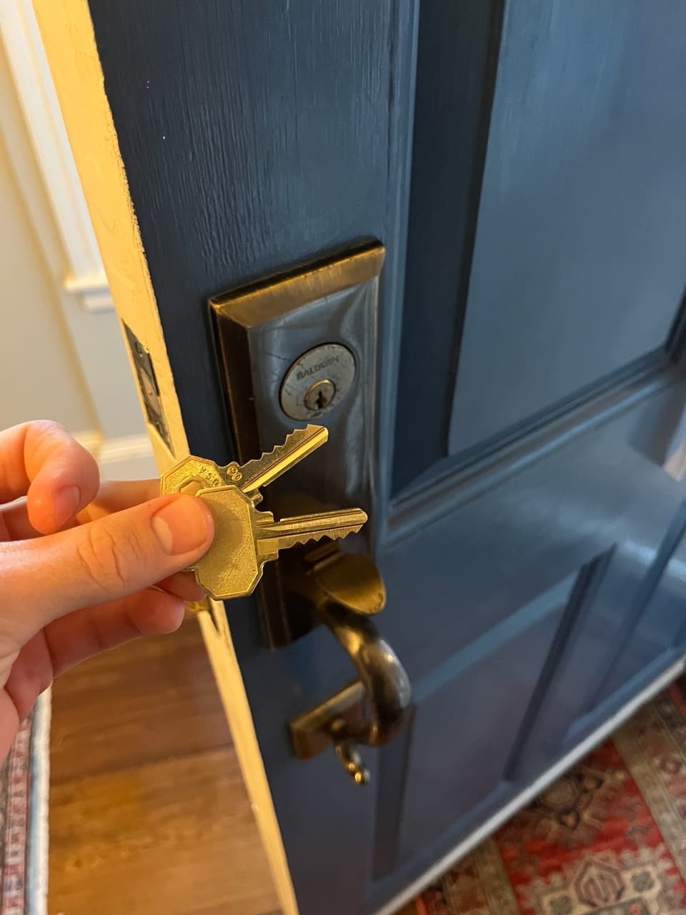 Hand holding two brass keys in front of a dark blue door with brass hardware.