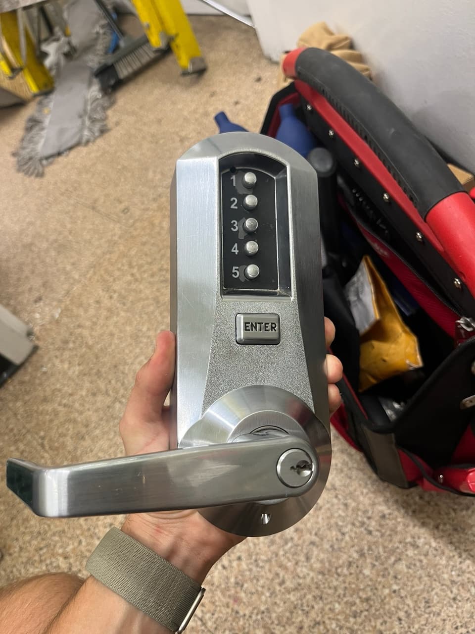 Hand holding a silver commercial door lock with a five-button keypad and enter button.