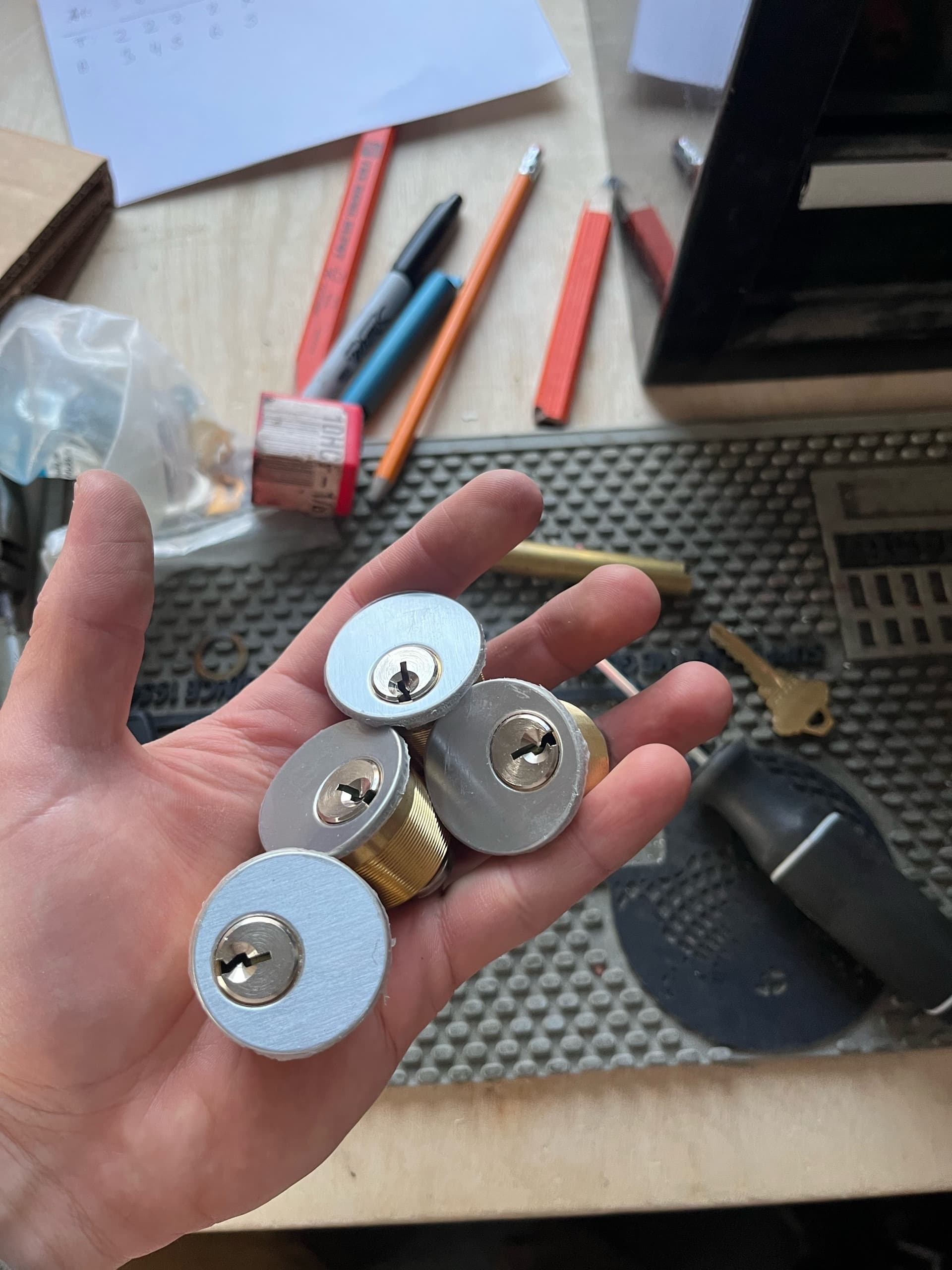 Hand holding four silver and brass mortise lock cylinders over a cluttered locksmith workbench.